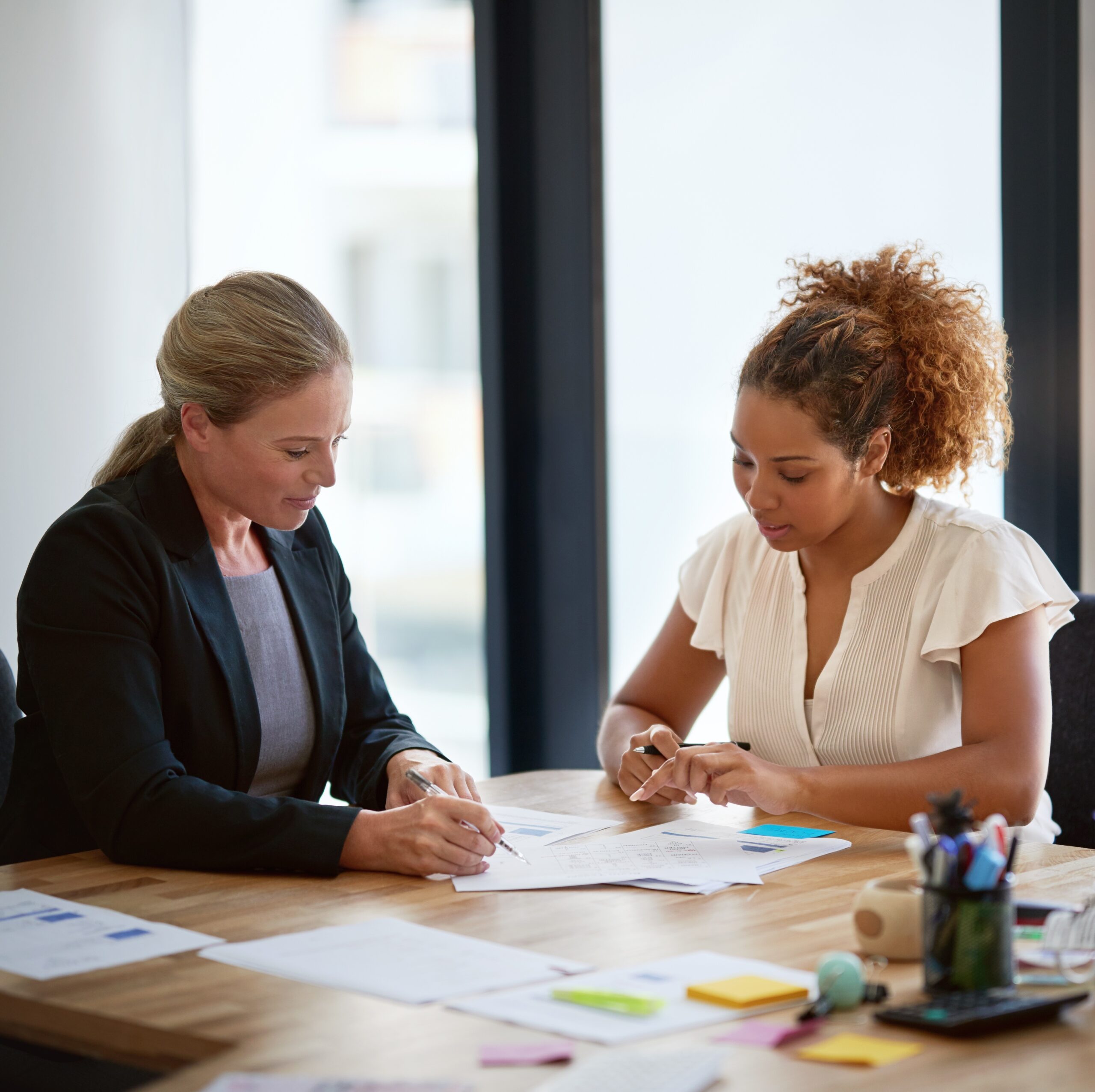 Two women collaborate at a desk in a professional setting. One takes notes while the other gestures toward the documents on the table.